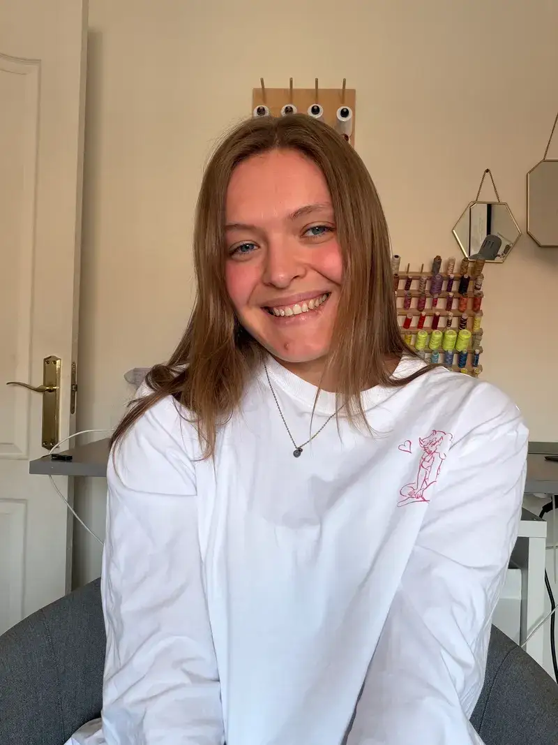 Victoria smiling in her studio, with a rack of coloured thread spools on the wall behind her